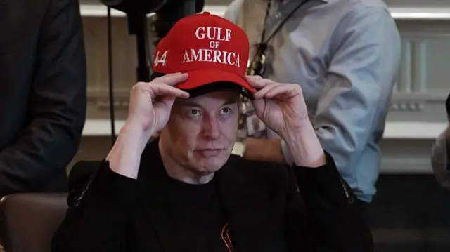 Elon Musk wearing red cap on top of another cap