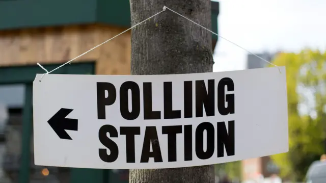 A white rectangular sign that reads polling on the first line, and station on the second. there is an arrow on the left pointing left. all font is in black. the sign is attached to string and pinned to the trunk of a tree outdoors.