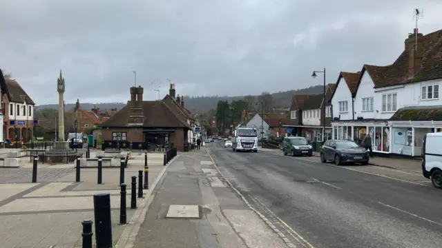 A picture of Wendover high street in Buckinghamshire. There is a road with shops alongside it and a lorry driving up the road towards the camera. On the left there is a clock tower.