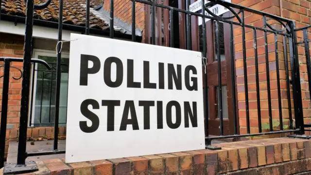 White cardboard reading polling station in black print ink, a red-brick building with a brown door behind the black railing on which the sign is hung