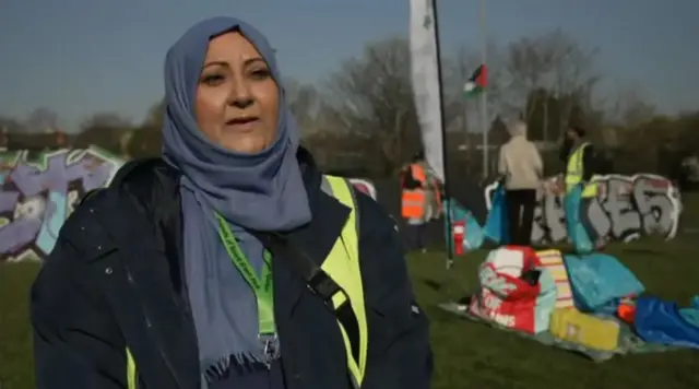 A woman with a blue headscarf and black coat standing in a field with piles of rubbish behind her