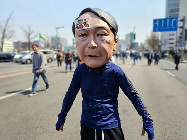 A person wearing a papier-mache mask of Yoon on their head.