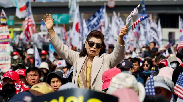 A woman frowning as she stands among a crowd, holding the South Korean and US flag.