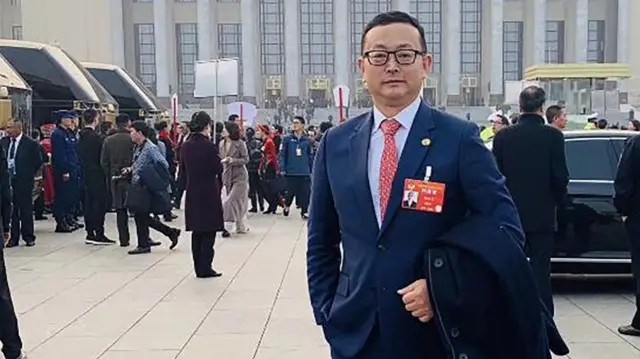 Yang Tengbo wearing a suit and glasses in front of a government building in Beijing during a CCP party gathering