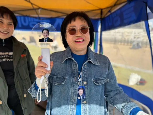 Seohyun Han, with short hair and sunglasses, wearing a denim jacket and holding a light stick with Lee Jae-myung's face printed on it