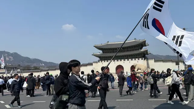A young man waving a giant South Korean flag. Around him are people walking around, and in the background is the gate to a Korean palace.