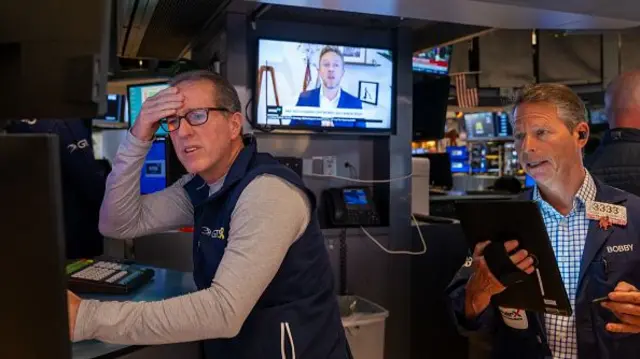 Traders work on the floor of the New York Stock Exchange. One man wearing spectacles has his right hand on his head while another man behind him holds an iPad device