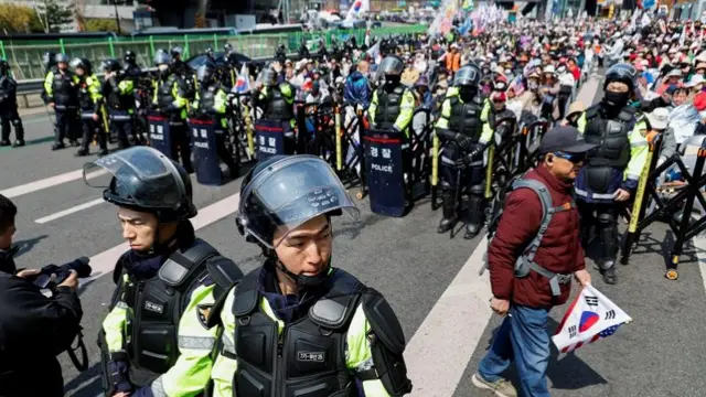 Security officers in helmets stand behind a barricade. Behind them are a crowd of protesters