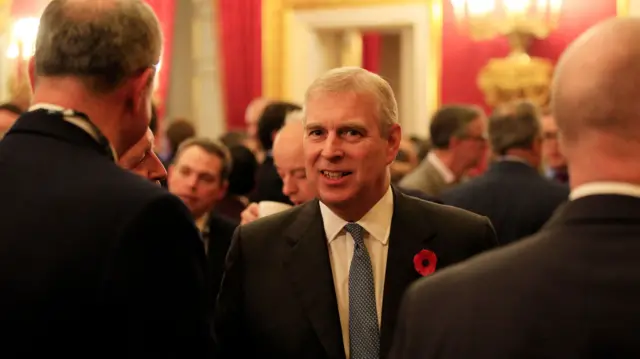 Prince Andrew stands facing the camera in a room of people. He wears a poppy