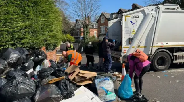 People standing next to a large pile of bin bags in the street with a white refuse lorry behind them