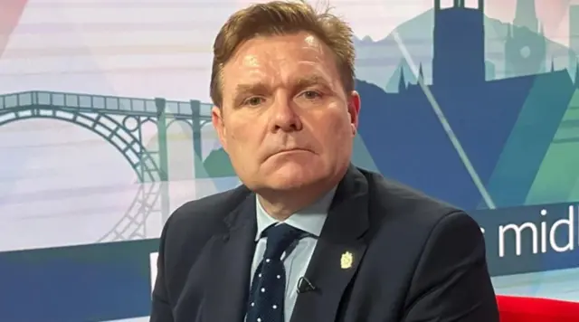 A man with ginger hair and a dark blue suit and tie sat on a red chair with pictures painted on a blue wall behind him