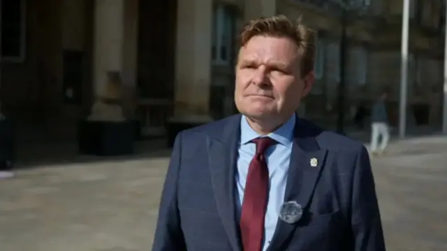 A man with brown hair standing outdoors in a navy suit and red tie