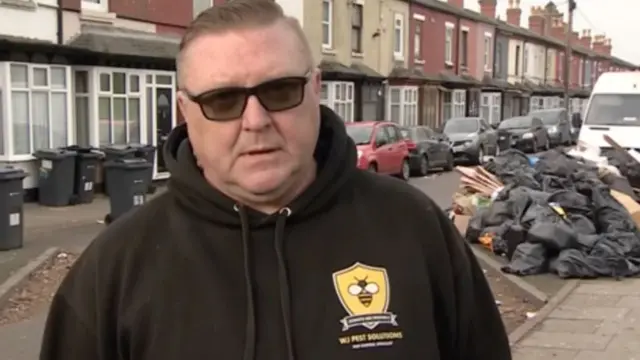 A man with short hair and sunglasses in a black fleece with an embroidered bee on it, standing in front of a street with lots of bin bags in it