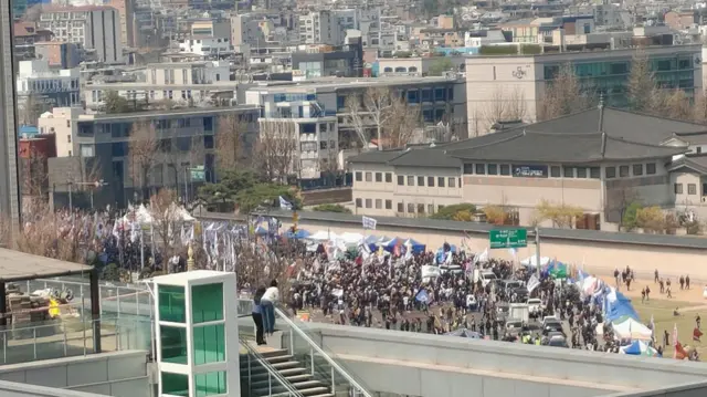 Wide shot of a crowd gathering on a field. In the background are Seoul's buildings.