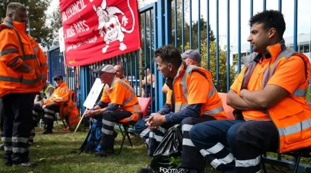 Men in orange reflective jackets sat on chairs in front of blue metal railings
