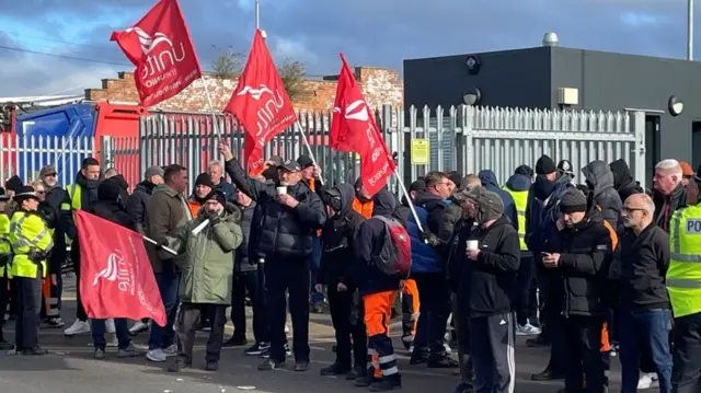 A gathering of men in dark clothing, holding red flags with the word "Unite" on them, with police officers in yellow reflective jackets on either side