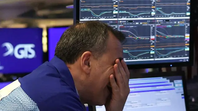 A trader hold his face in his hands as he reacts to slumping stock markets, reflected on a screen behind him.