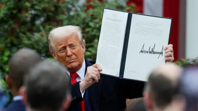 Donald Trump holds up a signed executive order. The backs of peoples heads are in the foreground facing the president - they have been blurred where he is in focus.