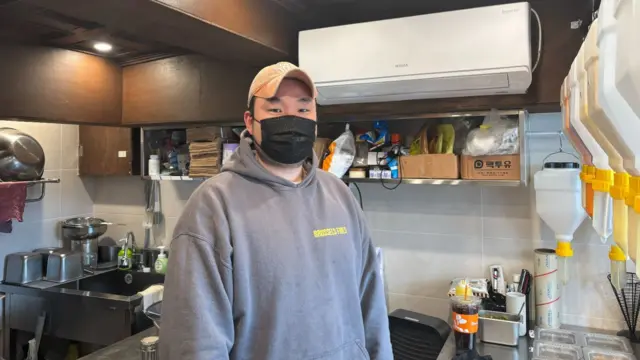 Choi Seung-yeol, wearing a grey hoody and black mask, looks at the camera in his kitchen