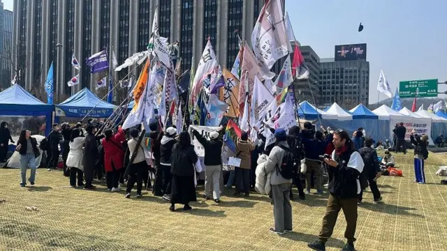 People gathering with giant flags in a field. Behind them are blue tents.