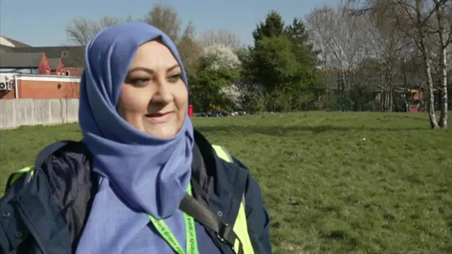Head and shoulders shot of a woman standing in a park, wearing a light blue top and headscarf