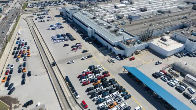 A drone view shows Stellantis's Chrysler Windsor Assembly facility in Windsor, Ontario, Canada. There is a large assembly facility surrounded by numerous parked cars and road on the left.