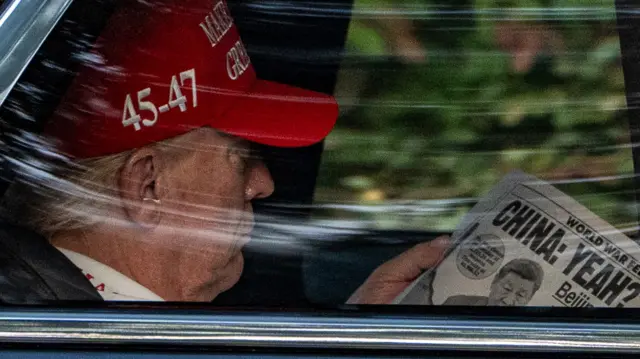 Donald Trump reads a double-page spread in the New York Post, headlined: "China: Yeah?" The president is wearing a red MAGA hat.