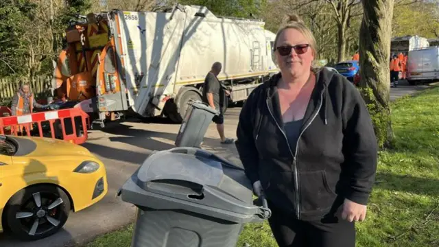 A woman in a black hoodie, holding the handle of a grey wheelie bin near a mobile collection truck.