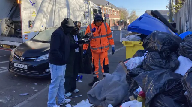 BBC radio presenter speaks to refuse worker stood in front of a pile of rubbish