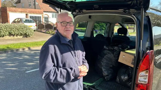 A man with grey hair and glasses in a blue fleece standing at the back of car with its boot open and bin bags in the back