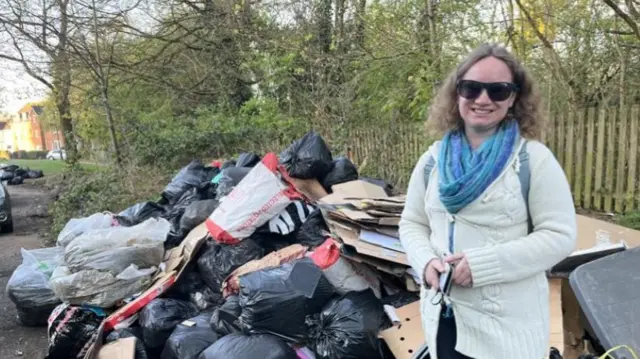 A woman with blonde hair, sunglasses and a white cardigan in front of a pile of bin bags