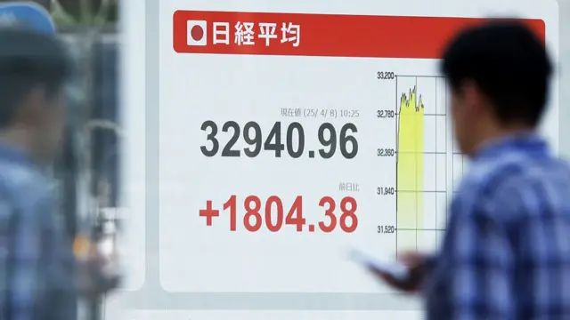 A man holding a phone looks at a digital screen showing the NIkkei 225 index