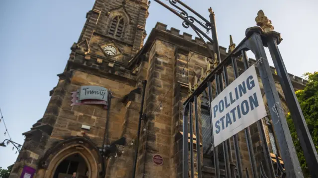 Tall church-like building seen in the background. A black ornate gate to the side has a white sign attached to it by string, reading: "Polling Station"