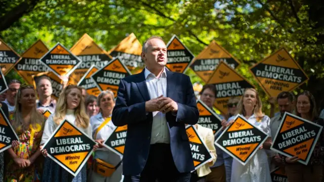 Ed Davey standing in front of a group of campaigners all holding up orange Liberal Democrat signs