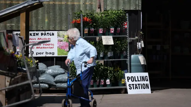 Woman using a walking aid stands in front of a shelf filled with plants. A sign reading "Polling Station" sits propped up against it