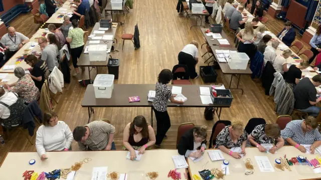 Seen from above, people sit at rows of tables arranged in a U shape, checking votes votes.