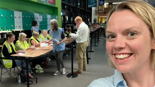 A selfie of Hannah Miller at the election count in Bristol. She is smiling at the camera. Behind her are people at a long table counting ballots.