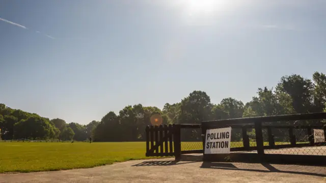 Sun shines in the sky over field. There's a gate in the foreground which has white sign with the words "Polling Station" in black
