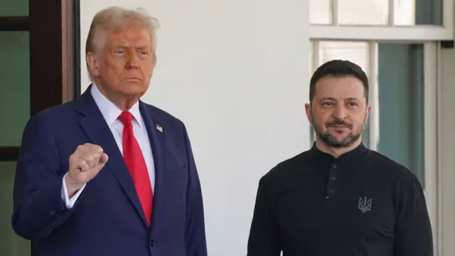 President Donald Trump stands next to President Volodymyr Zelensky. President Trump wears a blue suit with a red tie and President Zelensky wears a black button up top.
