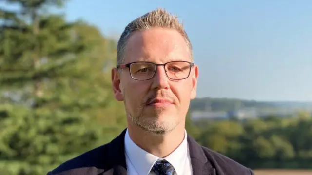 A man with short grey hair spiked up, a short grey beard and glasses stands outside looking at the camera. He is wearing a white shirt, a blue tie and a dark jacket.
