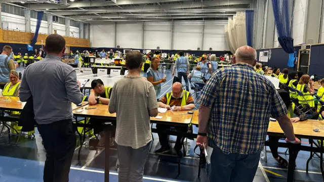 Three people are seen in the foreground facing away from the camera and towards rows of tables where people are counting votes.