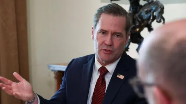 Mike Waltz speaks at a meeting at the White House. He is wearing a blue suit over a white shirt and a red tie.