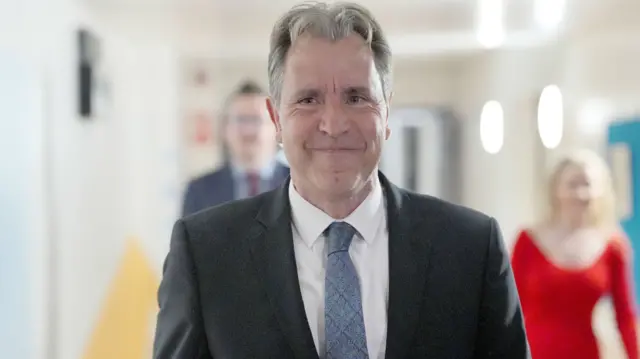 Dan Norris wearing a suit with a blue patterned tie with a tight lipped smile looking off camera