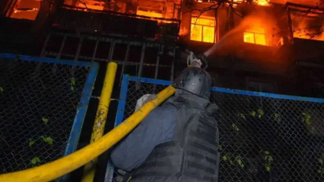 A firefighter holds a yellow hose to fire water at a burning building
