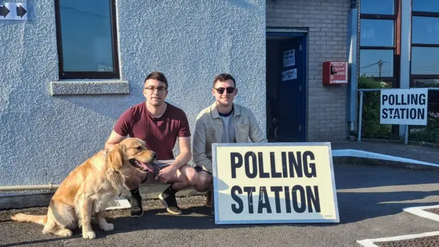 Two men crouch behind a 'Polling Station' sign with a Golden Retriever