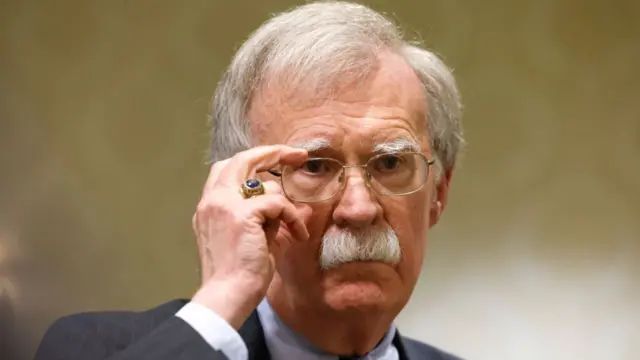 John Bolton, in a black striped suit with a large black ring on his finger, adjusts his glasses.