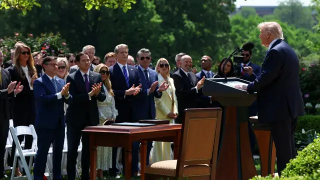 Donald Trump behind podium, speaking to group of officials, who are standing and clapping in the sunshine.
