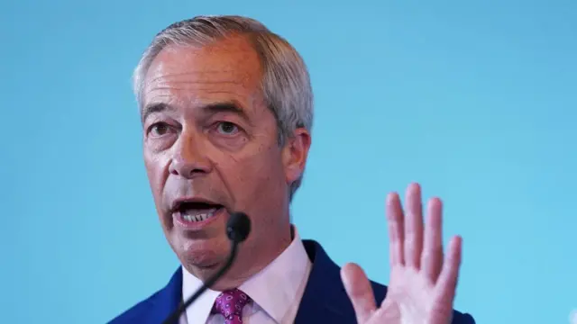 A file photo of Nigel Farage standing in front of a blue background and speaking into a microphone