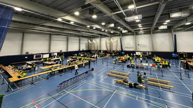 View from above over rows of tables where people are counting votes inside a sports hall with blue floors.