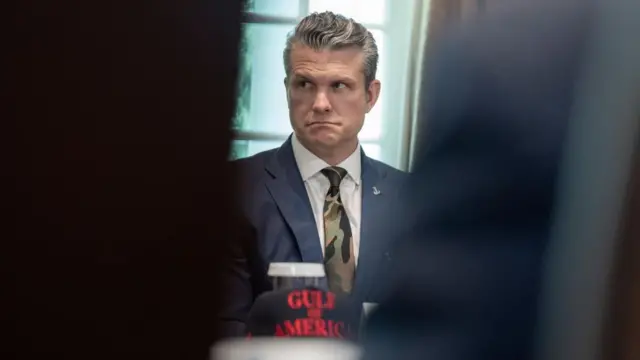 Hegseth, seated, is wearing a blue and black suit over a white shirt and a camouflage tie. A hat with the words Gulf of America is on the table in front of him.